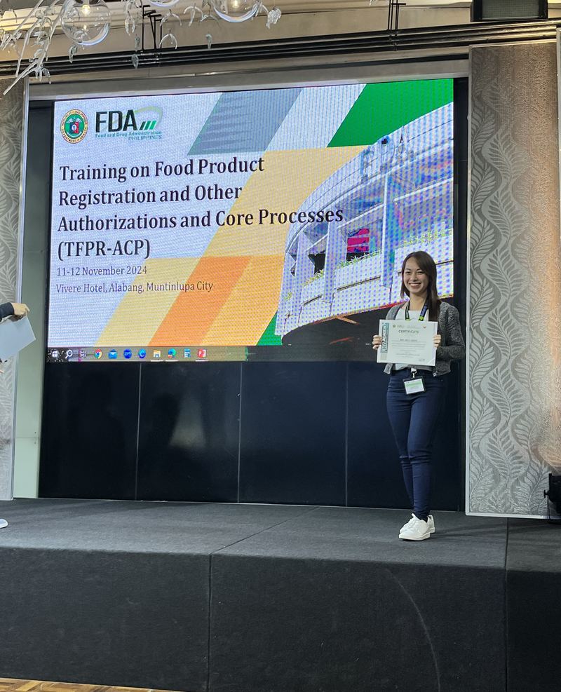 Mary Joice on stage holding her certificate at the FDA Training on Food Product Registration, November 2024